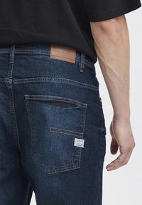 Tamno plavi traperice s smešanim kožnim etiketam, detaljno šivanje i bijela oznaka na stražnjem džepu. Klasičan dizajn s pet džepova.