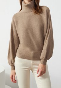Khaki ribbed turtleneck sweater with balloon sleeves, made from a soft knit material, paired with white trousers.