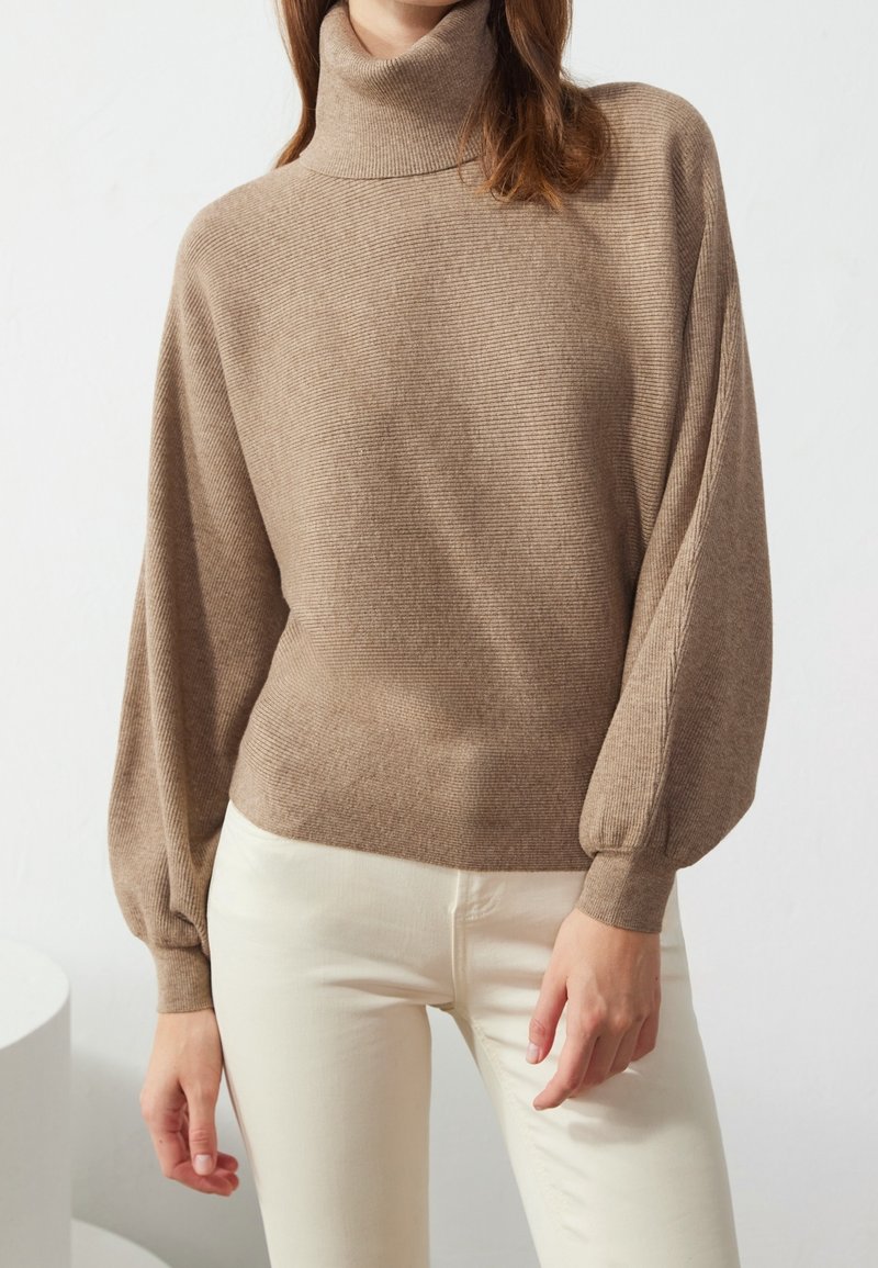 Khaki ribbed turtleneck sweater with balloon sleeves, made from a soft knit material, paired with white trousers.
