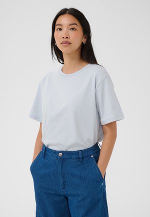 Femme aux longs cheveux foncés portant un t-shirt bleu clair oversize rentré dans un jean bleu taille haute, mains dans les poches sur un fond uni.