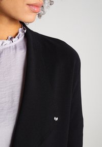 Schwarzer Cardigan aus weichem Stoff mit dezentem Glanz, versehen mit einem kleinen silbernen Schleifenakzent und zarten diagonalen Nähten.