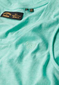 Canottiera turchese chiaro realizzata in tessuto morbido. Presenta un colletto arrotondato e il logo "Superdry" embossato vicino all'orlo.