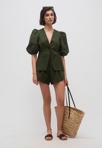 Kvinde iført olivengrøn top med pufærmer og matchende shorts, der holder en stor vævet stråtaske med sorte håndtag.