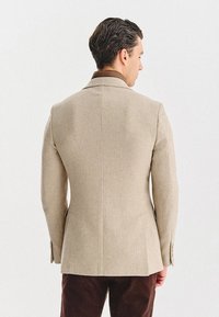 Mężczyzna odwrócony plecami, ubrany w beżową marynarkę w prążki, brązową koszulę i ciemnobrązowe spodnie, stojący na tle białego tła.