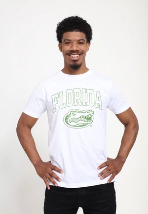 Lachende man met krullend haar die een wit T-shirt draagt met groene "FLORIDA" tekst en een alligatorlogo, staand met handen in de zij.