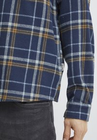 Camisa de franela en color navy con un estampado de cuadros en azul claro y marrón, con una abertura lateral y una etiqueta de la marca visible en el dobladillo.