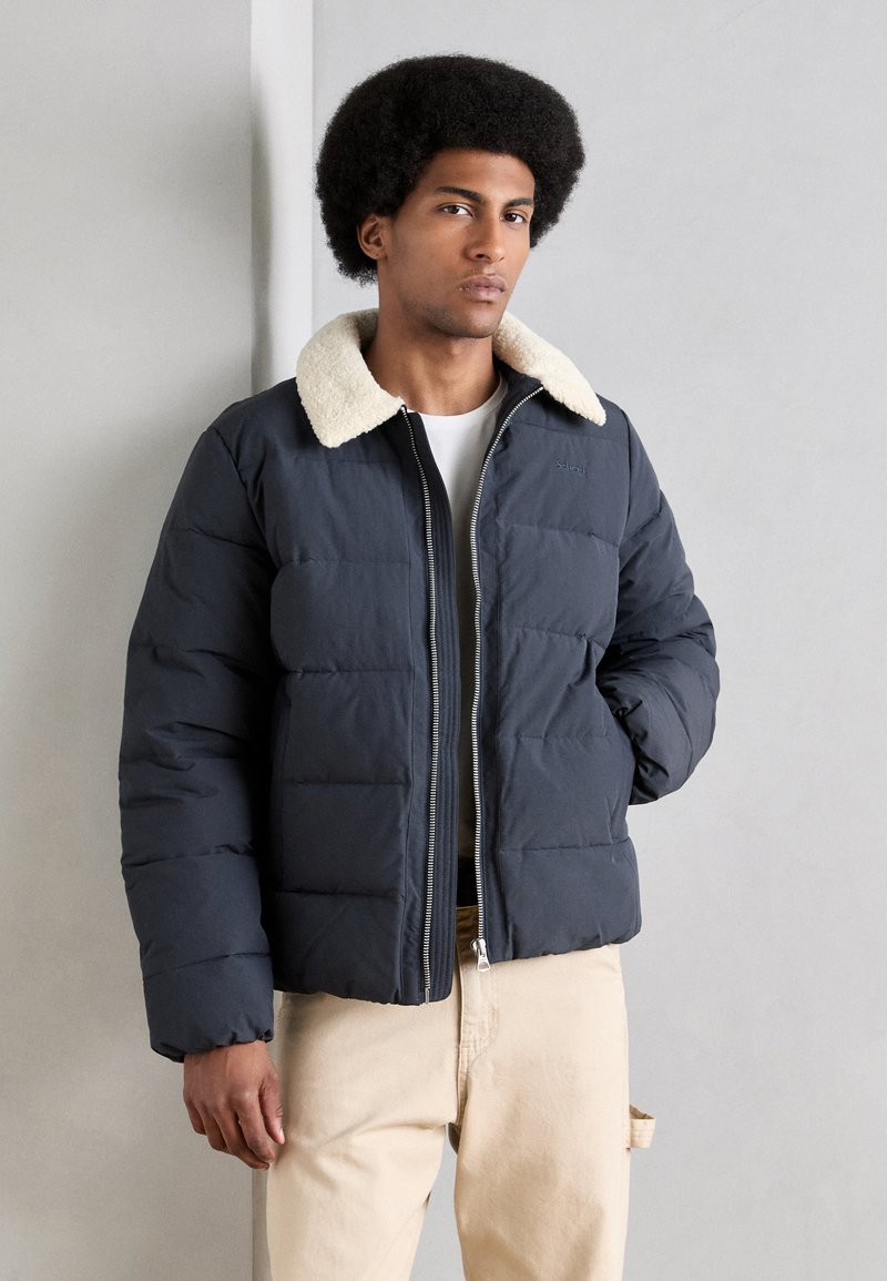 Pufferjakke i navy med en faux fur-krave, med lynlås og quiltet design. Båret over en hvid skjorte og khaki bukser.