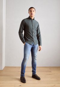 Dark green button-down shirt, blue jeans, and black loafers; model stands against a light gray wall with wooden floor.