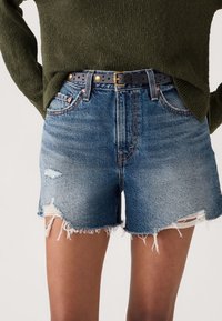 Personne portant un short en denim bleu usé avec des bords effilochés et une ceinture noire, associé à un pull en maille vert foncé.