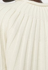 Saha beige-asutettu neulepusero, jossa on pystysuora ribbikuvio, pehmeä tekstuuri ja pyöreä pääntie. Ei näkyviä koristeita.