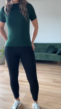 Top justo de mangas curtas verde escuro, combinado com leggings pretas. Ténis brancos com detalhes pretos. Piso de madeira e sofá verde ao fundo.
