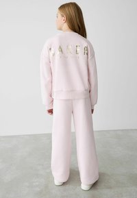 Ljusrosa sweatshirt och matchande byxor. Sweatshirten har "BAKER" i metallisk guldtext på baksidan. Mjuk tyg, avslappnad passform.