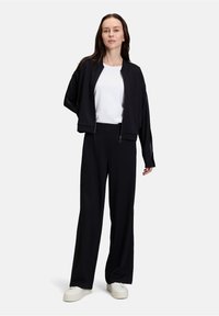 Veste bomber noire zippée sur un t-shirt blanc, associée à un pantalon large noir. Chaussures : baskets blanches. Tissu lisse, coupe décontractée.