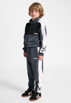 Blond włosów dziecko w czarno-szarym dresie Hummel z białymi paskami i czarnymi sportowymi butami, stoi i patrzy przed siebie na białym tle.