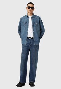 Denimskjorta och breda jeans i mörkblått, i kombination med en vit T-shirt. Anmärkningsvärda egenskaper inkluderar synlig sömnad och framfickor. Svarta skor.