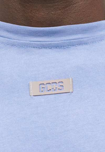 Λαιμός μπλουζάκια σε ανοιχτό γαλάζιο με μπεζ ετικέτα ραμμένη από κάτω, που δείχνει τα κομμένα γράμματα "GCDS".