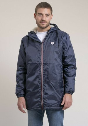 Homme portant une veste à capuche bleu marine avec doublure en polaire blanche, fermée par une fermeture éclair sur une chemise blanche, debout devant un fond uni.