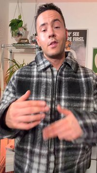 Young man with short dark hair wearing a black and white plaid zip-up jacket gesturing with blurred hands indoors.