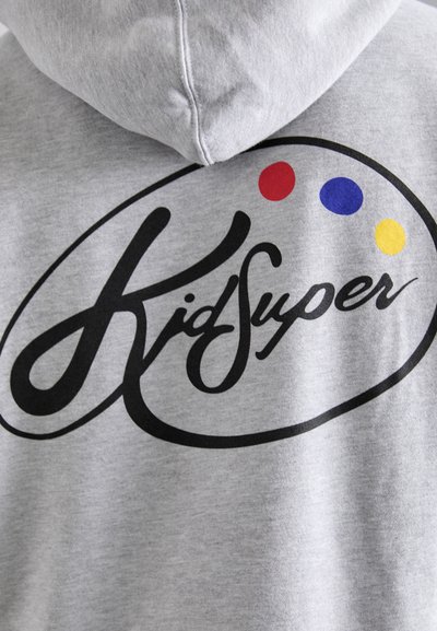 Siva kapuca z velikim črnim ovalnim logotipom s tekstom "KidSuper" v pisanem fontu, okrašena s rdečimi, modrimi in rumenimi pikami.
