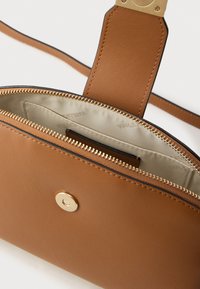 Barna bőr kézitáska sima textúrával, aranyszínű fémekkel, cipzáras záródással és bézs szövet bélésű belsővel, amely kártyatartó rekeszeket is tartalmaz.