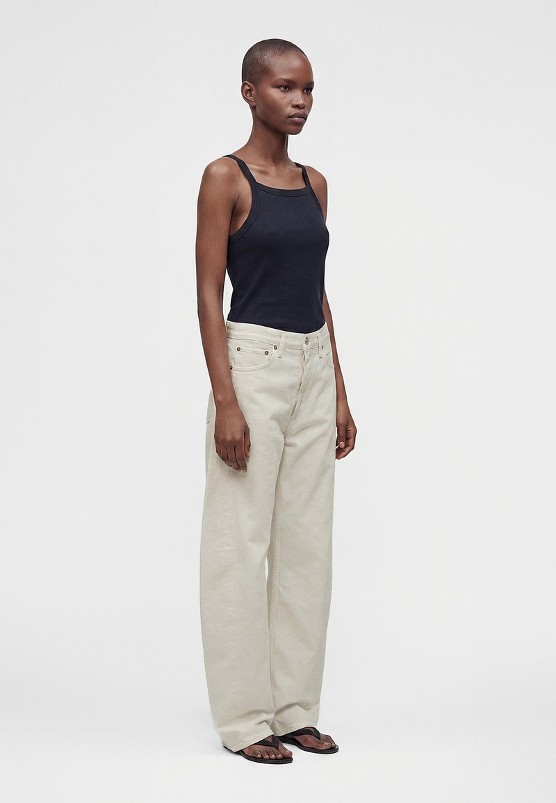 Femme debout portant un haut noir sans manches, un pantalon beige clair ample et des sandales noires sur un fond uni gris clair.