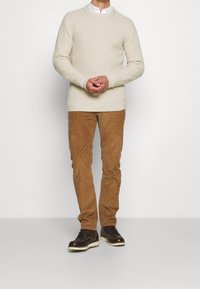 Lichtbeige kabelgebreide trui, bruine corduroy broek en donkerbruine veterlaarzen met witte zolen, geposeerd tegen een effen achtergrond.