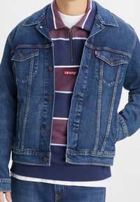 Veste en jean de couleur bleu foncé délavée, avec deux poches poitrine, des boutons en métal et un ourlet brut. Portée par-dessus un sweat à capuche zippé bleu marine à rayures.