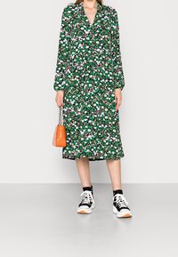 Vestito midi floreale con maniche lunghe, sfondo verde, fiori bianchi e viola, abbinato a sneaker in bianco e nero e una borsa arancione.