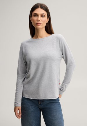 Vrouw die een lichtgrijs shirt met lange mouwen en blauwe spijkerbroek draagt, staande tegen een effen beige achtergrond.