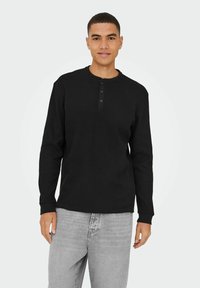 Sort langærmet henley skjorte med rund krave, tre-knap lukning og ribbet tekstur; parret med lysegrå jeans.