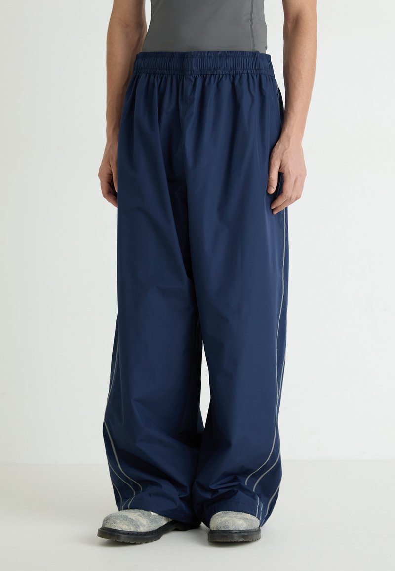 Navy waterproof pants with an elastic waistband, wide legs, and reflective side stitching. Worn over gray top and gray footwear.