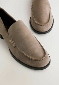 Mocassins en suede gris avec une texture lisse, un bout arrondi, des coutures décoratives et un petit talon en caoutchouc noir. Deux chaussures présentées de dessus.