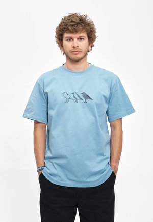 Jonge man met krullend haar die een lichtblauw T-shirt draagt met drie verschillende vogel silhouetten gedrukt op de borst.