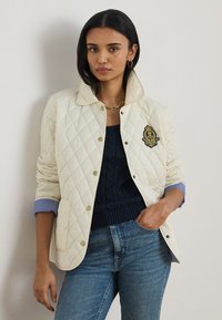 Femme portant une veste matelassée crème avec un écusson, un pull marine, des poignets de chemise rayés, un collier chaîne doré et un jean bleu de lavage moyen.