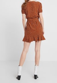 Robe marron à pois avec des manches courtes, une taille cintrée, un ourlet évasé et des détails en volants. Associée à des bottines blanches à talons.