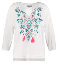Blouse blanche à manches longues avec broderie géométrique colorée et deux pompons roses suspendus à l'encolure.