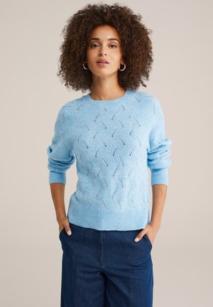 Pull en maille bleu clair avec un motif ondulé, des poignets et un ourlet côtelés. Le modèle le porte avec un pantalon en denim bleu foncé décontracté.