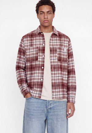Jeune homme portant une chemise à carreaux rouge et blanche boutonnée par-dessus un t-shirt beige avec un jean bleu clair, debout devant un fond blanc uni.