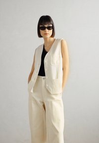 Crèmekleurige vest met knopen aan de voorkant en twee zakken, gedragen over een zwarte top en gecombineerd met wijd uitlopende crème broek. Model draagt een zwart zonnebril.