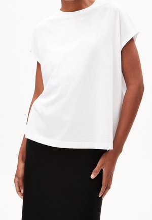 Nainen pukeutuneena yksinkertaiseen valkoiseen lyhythihaiseen toppiin ja mustaan ribbihameeseen, seisomassa yhdellä kädellä sivulla lepäämässä.