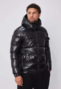 Veste noire matelassée avec finition brillante, col montant, fermeture éclair frontale et deux poches latérales. Présente un design matelassé avec des coutures apparentes.