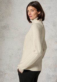 Cremfarbener Rollkragenpullover aus weichem Strick, mit gerippten Ärmeln und Saum. Getragen zu schwarzen Hosen, vor einem strukturierten neutralen Hintergrund präsentiert.