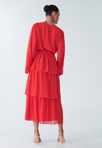 Abito in chiffon rosso a strati con maniche lunghe, vita elastica e fondo fluente. Indossato con sandali trasparenti con tacco.