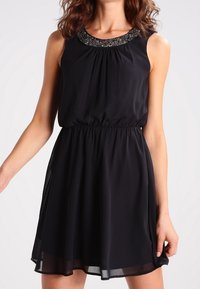 Robe noire sans manches avec une taille froncée, une jupe fluide et un décolleté orné de détails perlés. Confectionnée en tissu léger et transparent.