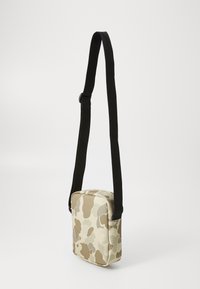 Sac bandoulière en tissu beige avec un motif camouflage. Il dispose d'une bandoulière noire réglable et d'une forme rectangulaire.