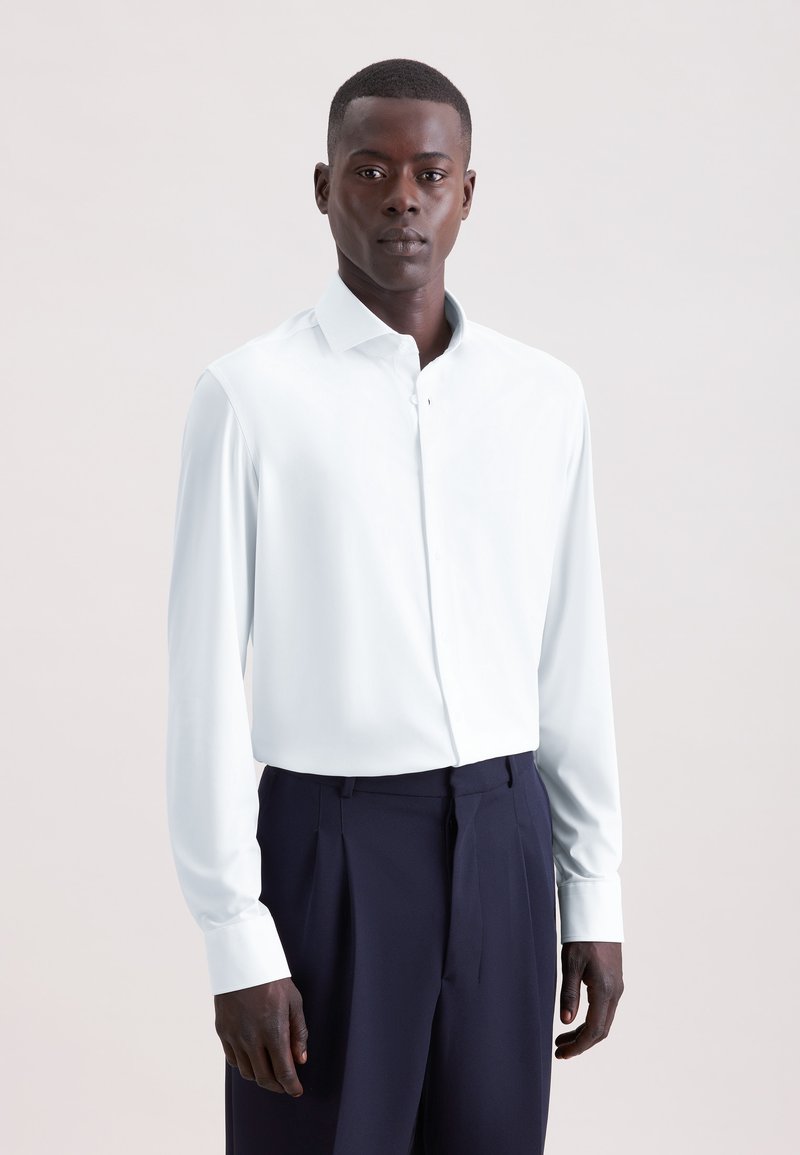 Homme portant une chemise blanche boutonnée et un pantalon bleu marine foncé, debout devant un fond clair uni.