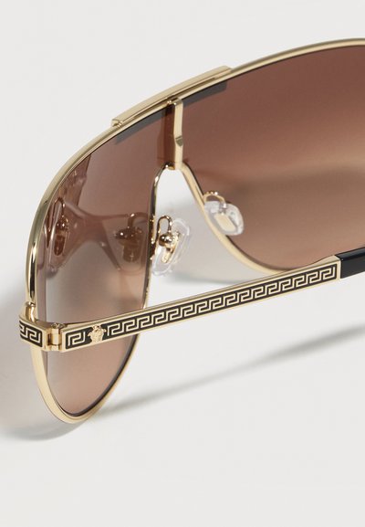 Lunettes de soleil à monture dorée avec des verres dégradés marron et des branches noires ornées d'un motif clé grec doré et d'un emblème Méduse.