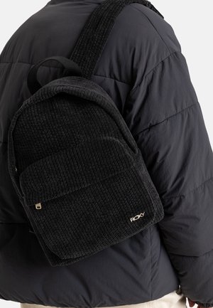 Zwarte corduroy rugzak met een frontzak en stoffen schouderbanden, gedragen door een persoon in een donkere pufferjack. Gouden logo accent.