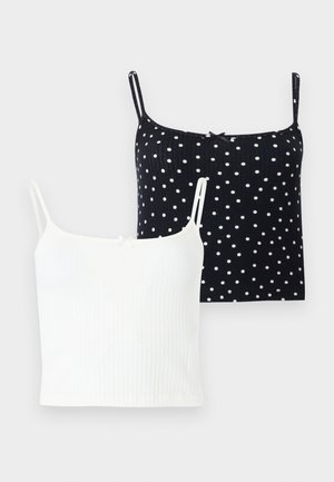 Jeune femme aux cheveux bouclés portant des lunettes de soleil rondes, un top court blanc sans manches, un jean bleu ample retroussé aux chevilles et des tongs noires.