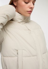 Beżowa kurtka puchowa z wysokim kołnierzem, pikowanym wzorem i boczną kieszenią z klapką. Gładki, wyścielany materiał poprawia izolację.